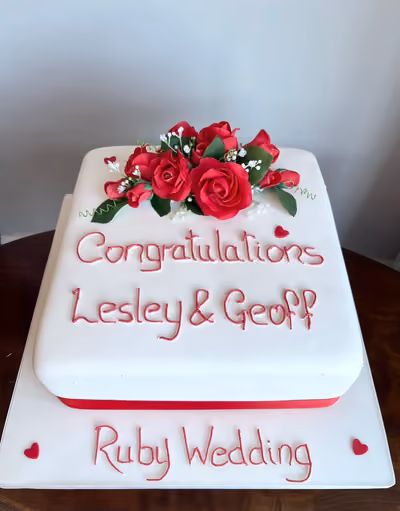 A square, two-tier white ruby wedding anniversary cake, decorated with a cluster of red sugar roses and small white flowers on top, a red ribbon around the base of the top tier, and red piped lettering that reads 'Congratulations Lesley & Geoff Ruby Wedding' with small red heart accents.