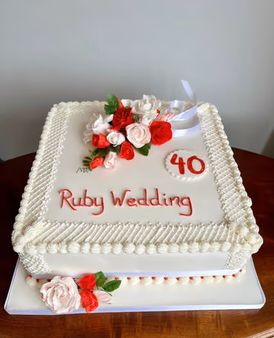 A two-tier square ruby wedding anniversary cake with white frosting, decorated with red and white sugar flowers, green leaves, and white ribbons. The words "Ruby Wedding" are written in red icing, and a circular plaque with the number "40" in red is also on the top tier.