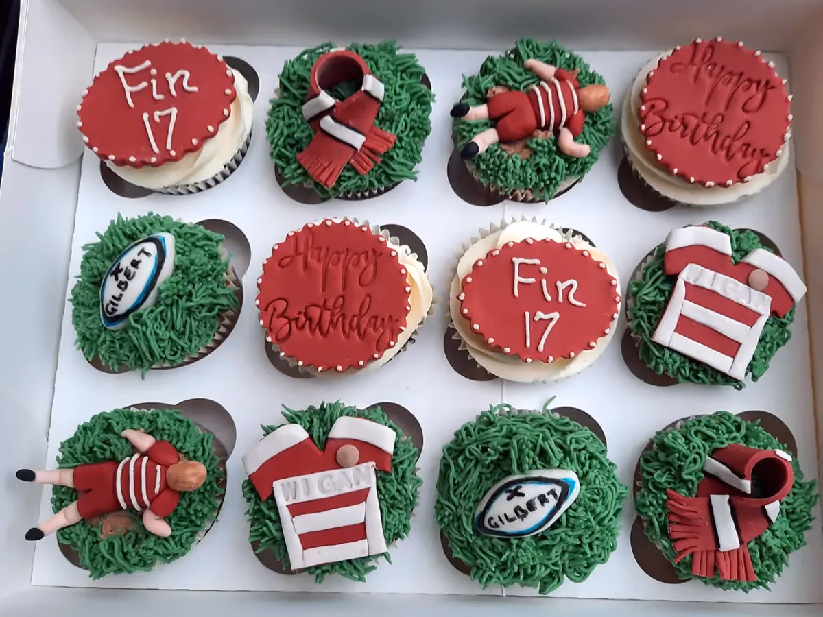A dozen rugby-themed birthday cupcakes in a white box, featuring green grass frosting, red and white decorations including rugby jerseys, player figures, rugby balls, scarves, and messages 'Fin 17' and 'Happy Birthday'.