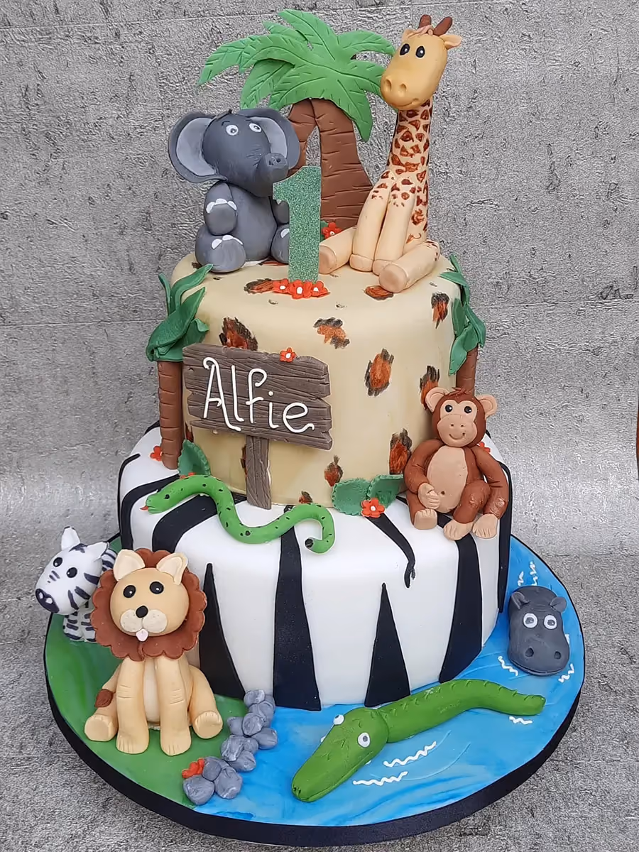 A two-tier safari-themed first birthday cake decorated with fondant animal figures including an elephant, giraffe, monkey, lion, zebra, hippo, crocodile, and snake. The top tier features a leopard print, a palm tree, and the number '1', while the bottom tier has a zebra stripe pattern with a water feature and a sign reading 'Alfie'.