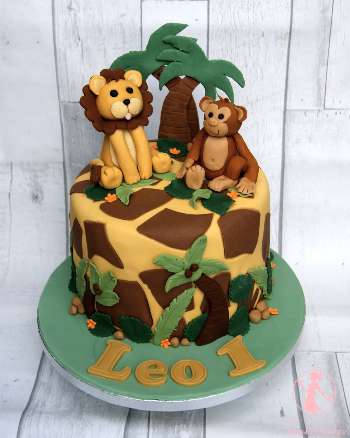 A single-tier safari-themed first birthday cake with a yellow and brown giraffe print pattern. On top are fondant figures of a lion and a monkey, accompanied by green palm trees and leaves. The green cake board features gold letters spelling 'Leo 1'.