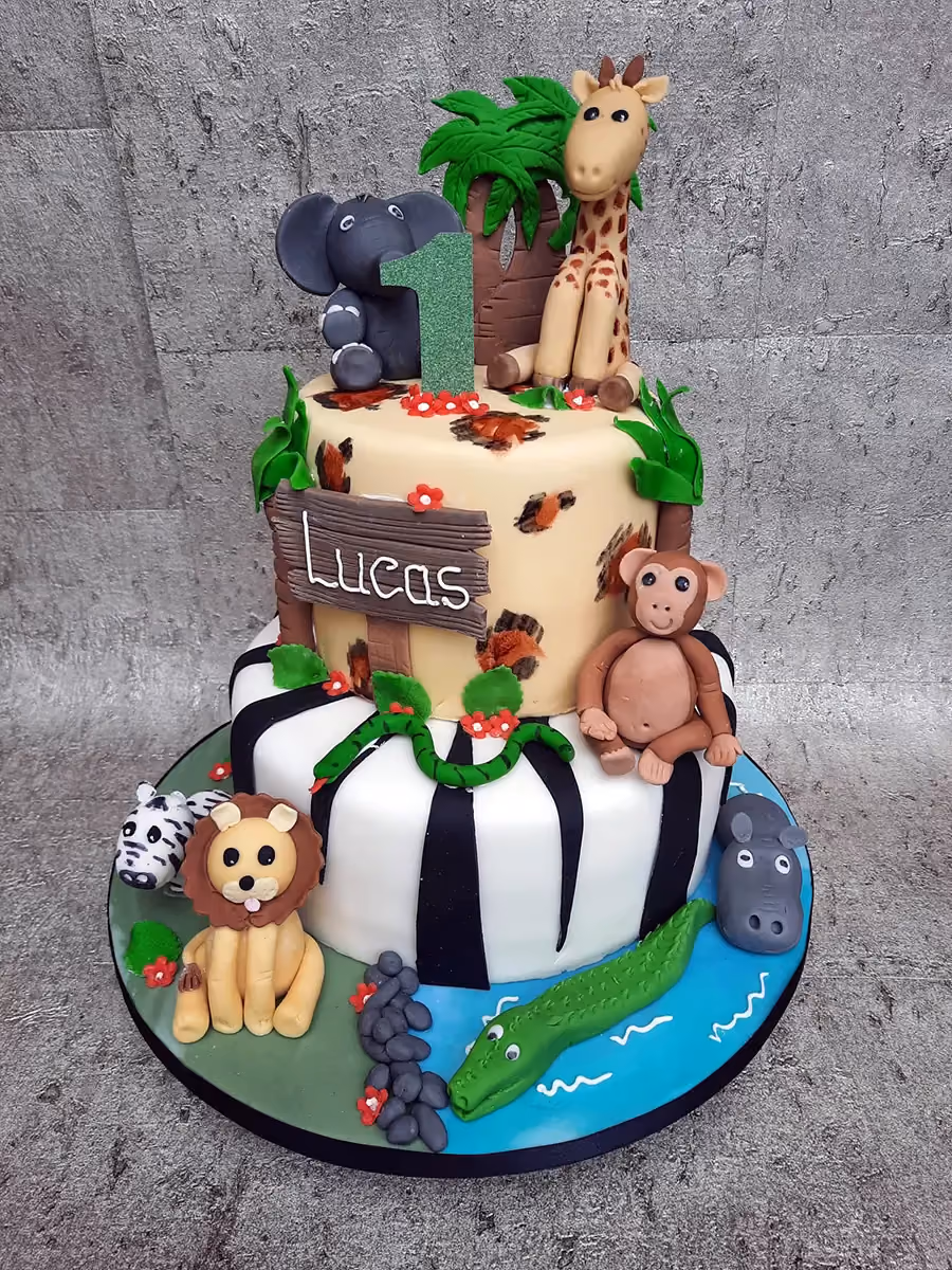 A vibrant two-tier safari-themed birthday cake for a first birthday, featuring numerous edible animal figurines. The top tier includes an elephant, a giraffe, a monkey, a green glittery number '1', and a 'Lucas' sign, surrounded by jungle foliage. The bottom tier has black and white zebra stripes, and the cake board is decorated with a zebra, a lion, a hippo, and a crocodile in a blue water pond.