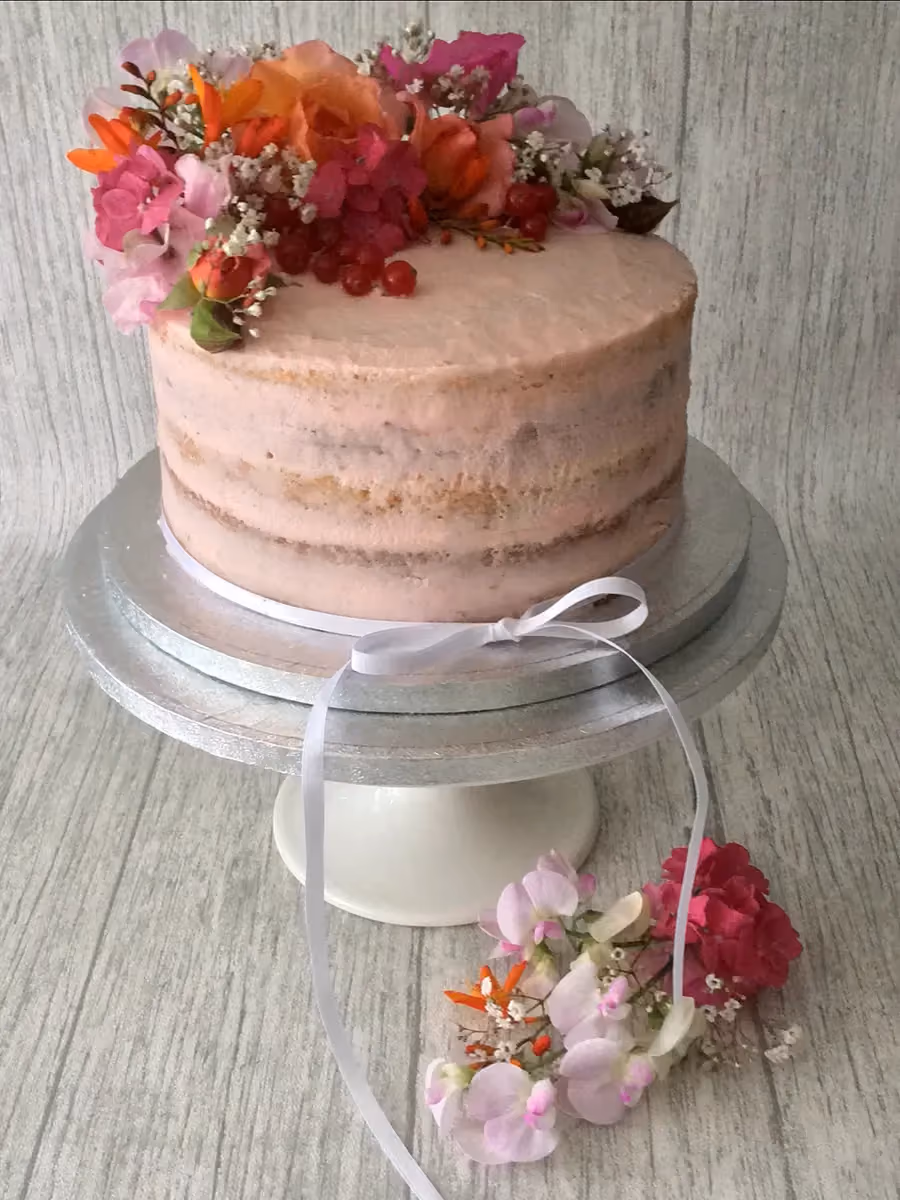 A single-tier semi-naked cake with light pink frosting, lavishly decorated with a bouquet of fresh pink, orange, fuchsia, and white flowers, and red currants on top. A white ribbon is tied around the cake board, with a matching small floral arrangement on the wooden surface.
