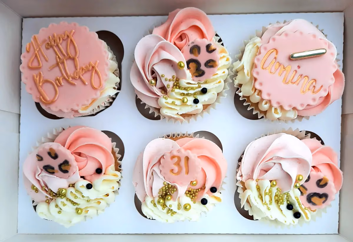 Six cupcakes arranged in a white box, each decorated with pink and white frosting, gold sprinkles, and black pearls. Two cupcakes have pink fondant toppers with gold text, one reading 'Happy Birthday' and another 'Amina', while a third has the number '31'. The remaining three cupcakes feature pink leopard print fondant accents.