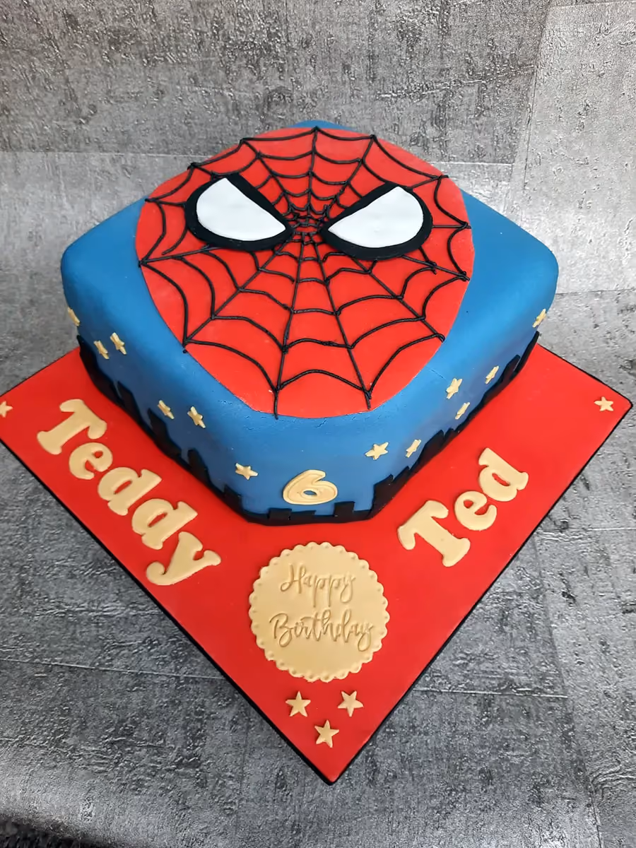 A square birthday cake featuring a blue base and a red Spiderman face on top with black web lines and white eyes. The cake is decorated with gold stars and a gold number 6, and sits on a red cake board with 'Teddy Ted' written in gold letters and a gold 'Happy Birthday' plaque.