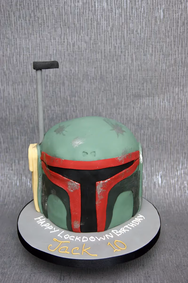 A Star Wars Boba Fett helmet-shaped birthday cake, featuring a distressed green, red, and black design, sitting on a grey cake board that reads 'HAPPY LOCKDOWN BIRTHDAY Jack 10' in white and yellow icing.