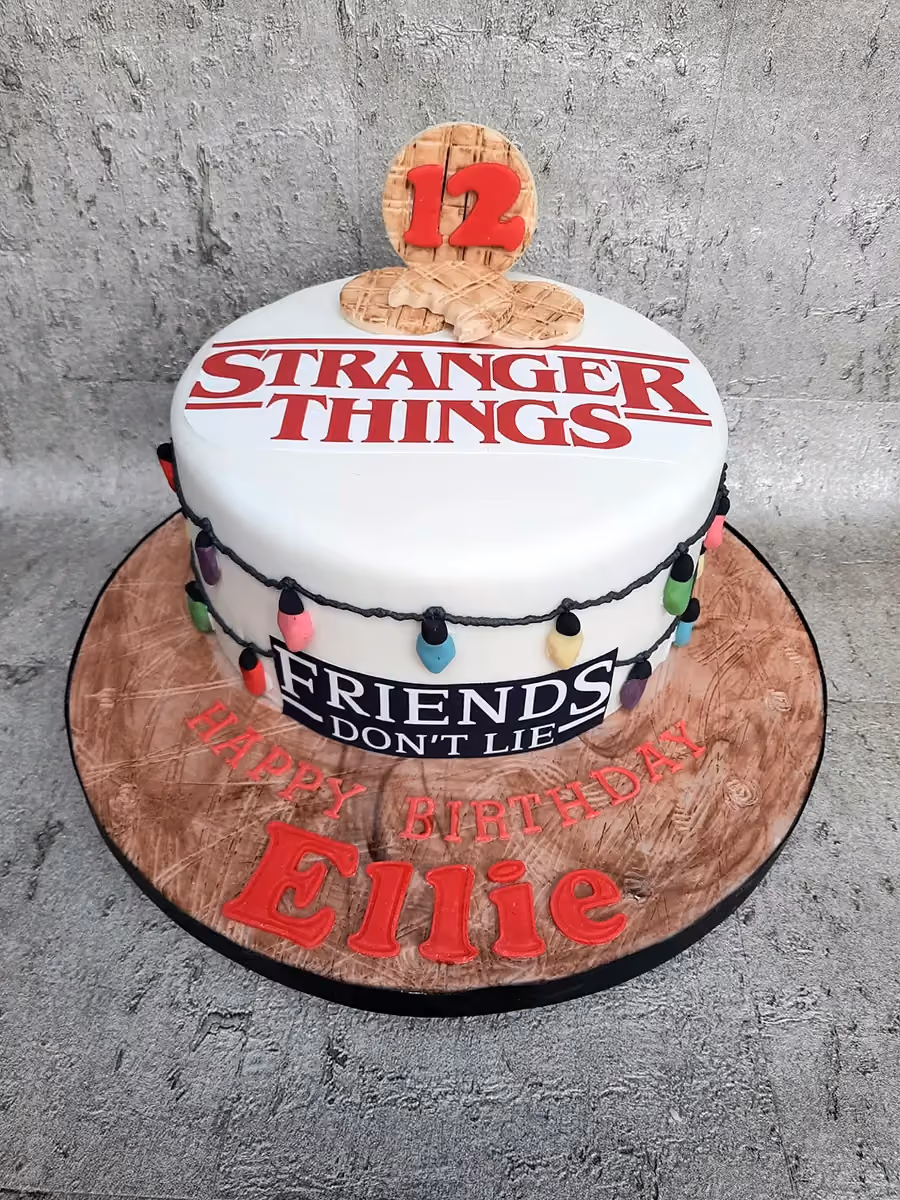 A white round Stranger Things themed birthday cake for Ellie's 12th birthday, featuring the show's logo, a string of colorful lights, and waffle decorations, with 'Happy Birthday Ellie' written in red on a wood-textured cake board.