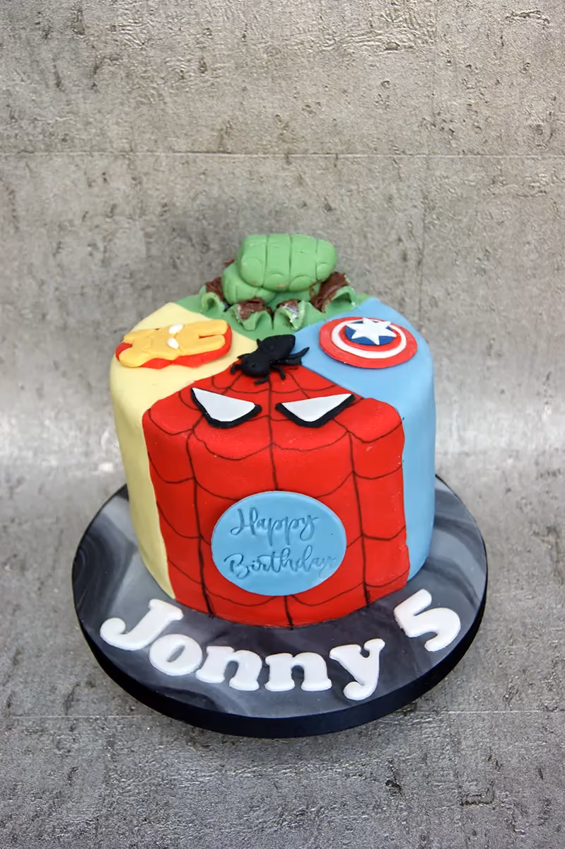 A round birthday cake decorated with four superhero themes: Hulk, Iron Man, Spider-Man, and Captain America. The cake has a light blue circle on the Spider-Man section with 'Happy Birthday' written on it, and the name 'Jonny' and the number '5' are spelled out in white letters on the black and grey marbled cake board.