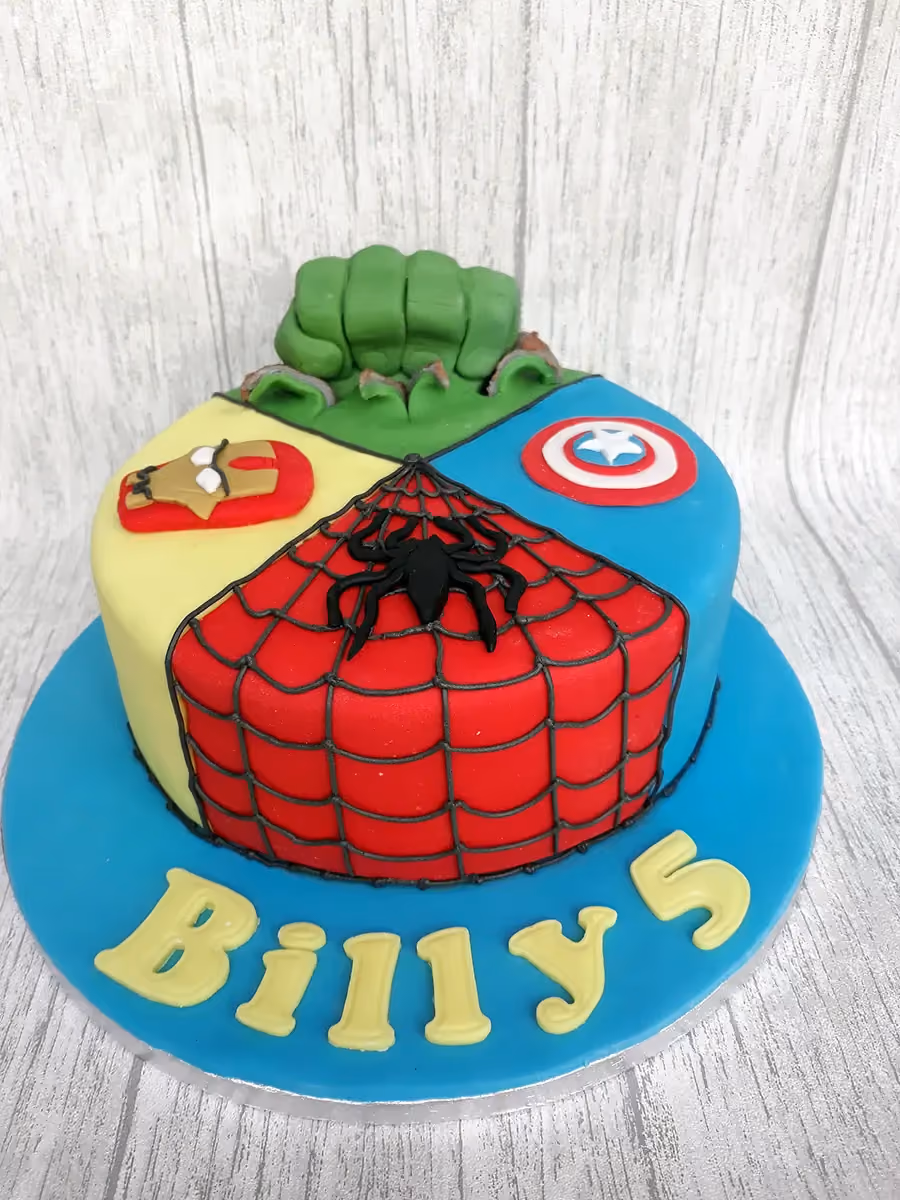 A round superhero-themed cake divided into four sections featuring fondant decorations of Hulk's fist, Iron Man's mask, Captain America's shield, and Spider-Man's web with a black spider, with 'Billy 5' written in yellow on the blue cake board.