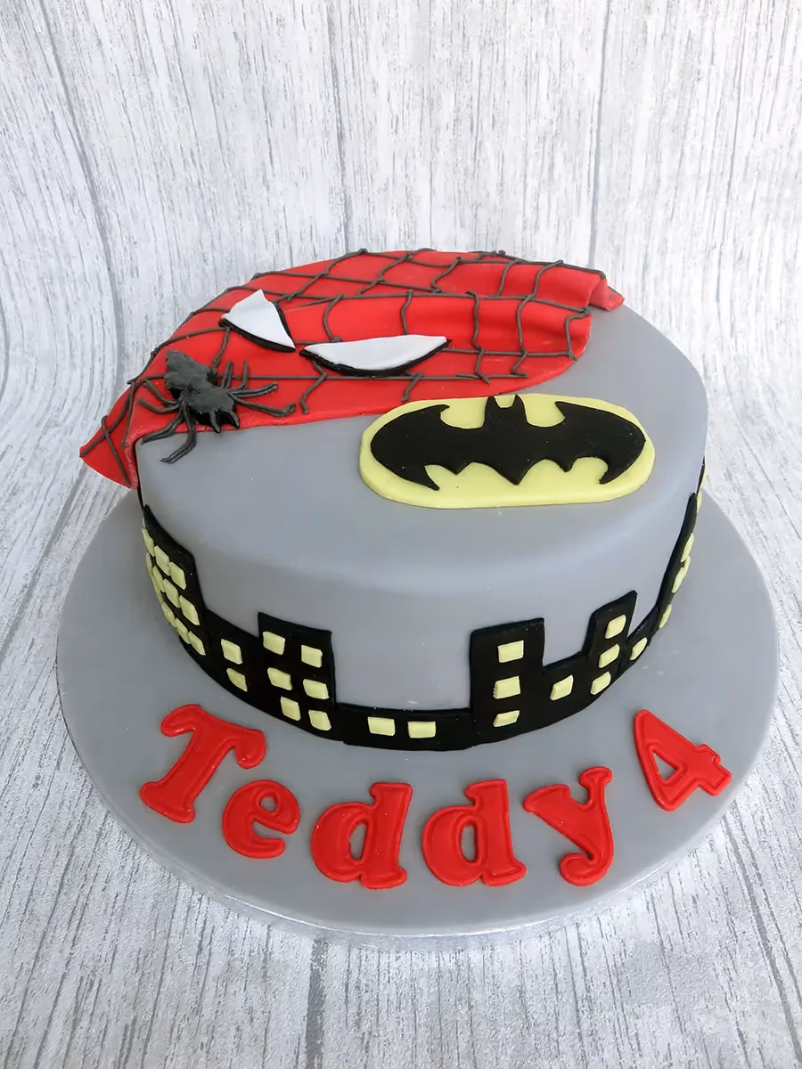 A single-tier grey fondant birthday cake featuring a red Spiderman mask, a black and yellow Batman logo, a black city skyline around the base, and the name 'Teddy 4' in red letters on the cake board.