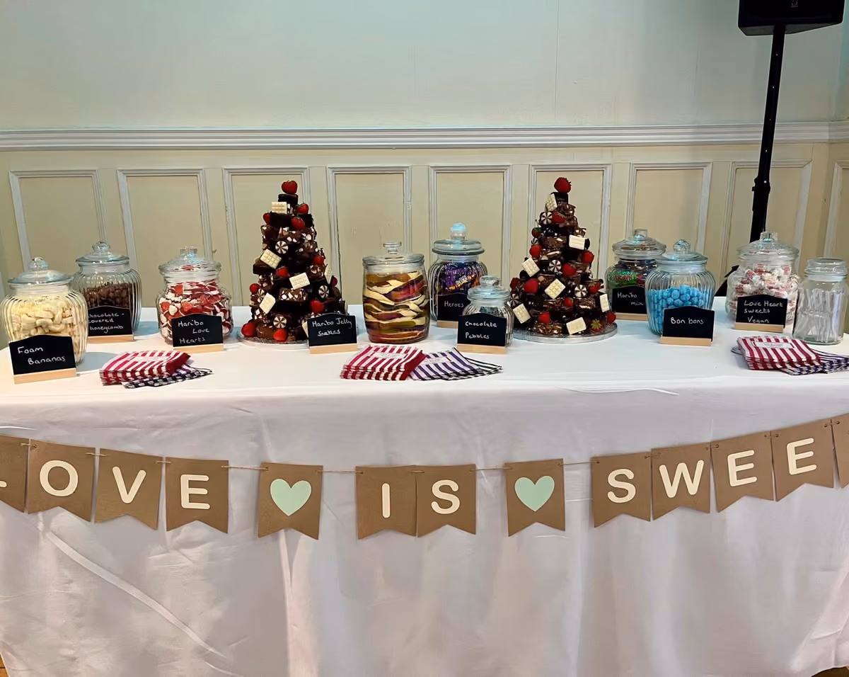 A sweet table featuring various glass jars filled with different candies and two chocolate brownie trees adorned with strawberries and white chocolate squares, with a 'Love is Sweet' banner hanging below.