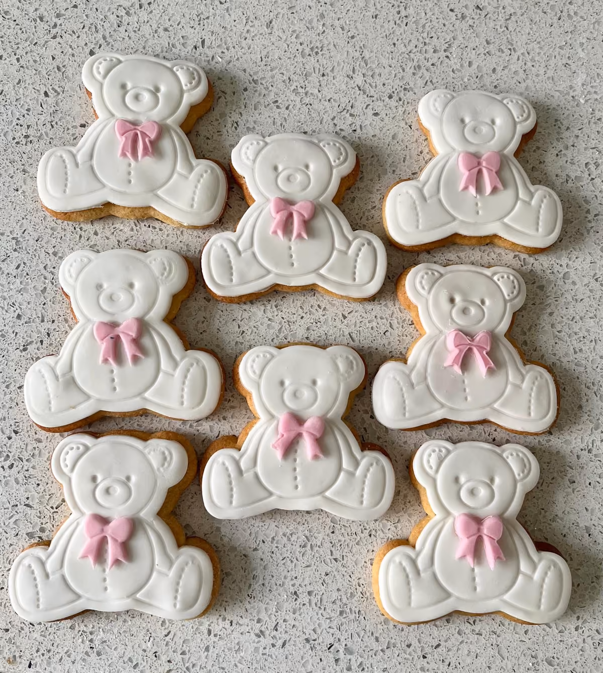 Nine white teddy bear shaped sugar cookies, each decorated with a small pink bow, laid out on a speckled light grey countertop.