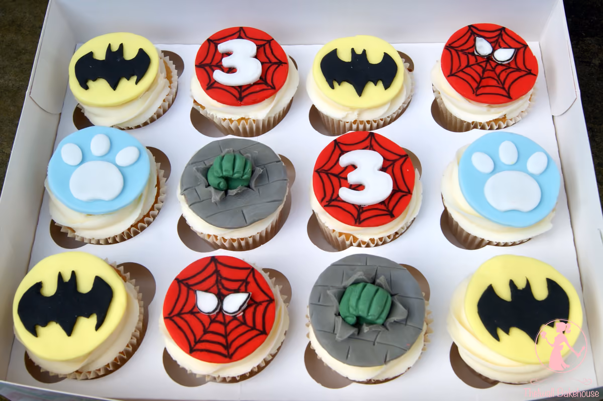 A white box containing 12 themed cupcakes decorated with yellow Batman logos, red Spider-Man webs (some with a white number 3), grey and green Hulk fists, and light blue paw prints.