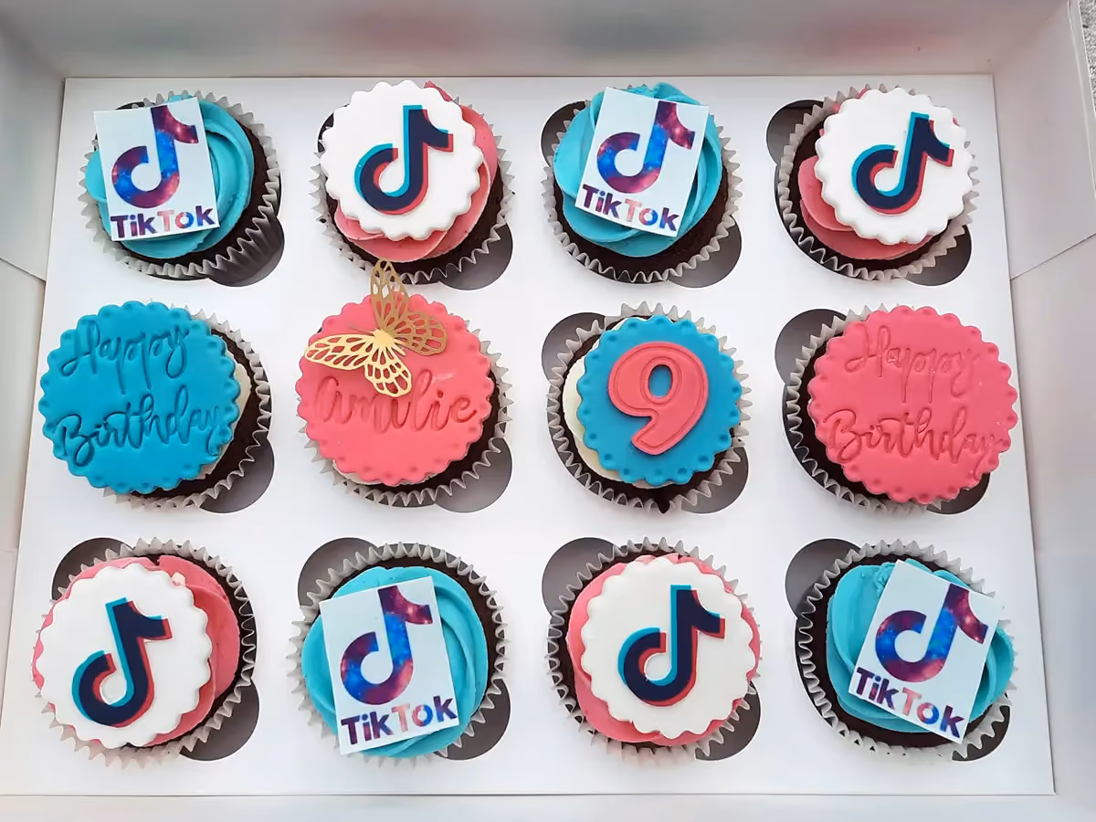 A white box containing twelve chocolate cupcakes, each topped with either pink or teal frosting and decorative fondant toppers. The toppers include the TikTok logo, 'Happy Birthday' text, the name 'Amelie' with a gold butterfly, and the number '9'.