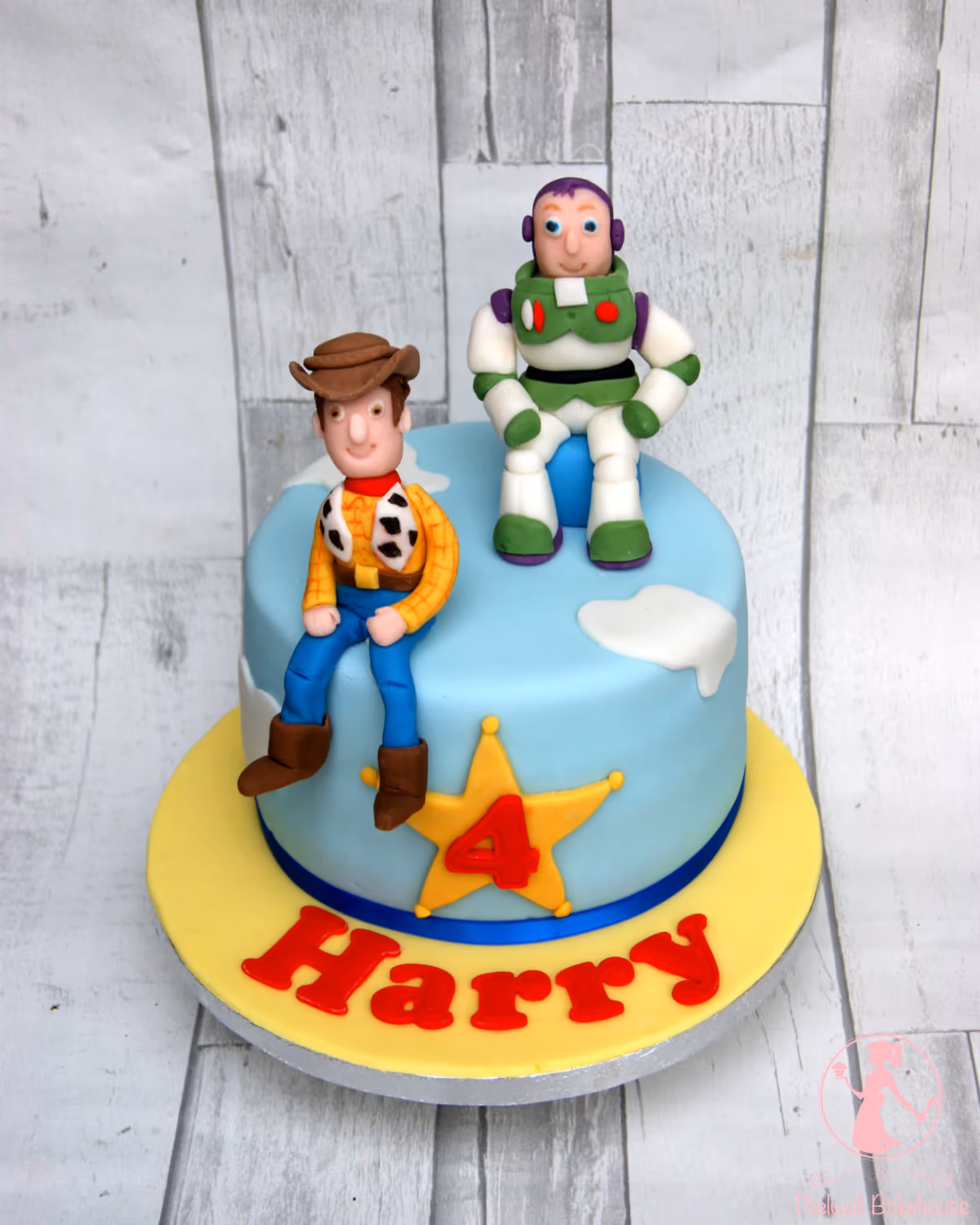 A round blue birthday cake decorated with fondant figures of Toy Story characters Woody and Buzz Lightyear, white cloud accents, a yellow sheriff's star with the number 4, and the name Harry spelled out in red letters on the yellow cake board.