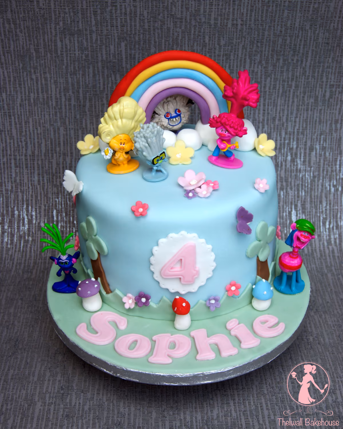 A light blue single-tier Trolls themed birthday cake for Sophie's 4th birthday, decorated with Trolls figures, a rainbow, flowers, mushrooms, and trees, with the name 'Sophie' on the base.