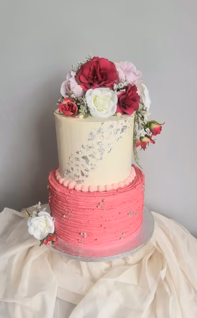 A two-tier celebration cake featuring a cream top tier with silver leaf accents and a textured bright pink bottom tier with silver sprinkles. The cake is adorned with a bouquet of pink, white, and burgundy artificial roses on top, and a small white rose with pink buds at its base.