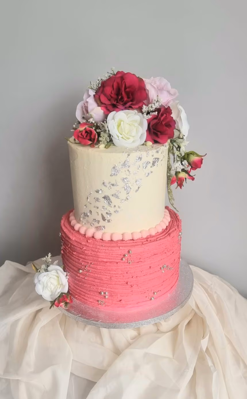 A two-tier celebration cake featuring a cream top tier with silver leaf accents and a textured bright pink bottom tier with silver sprinkles. The cake is adorned with a bouquet of pink, white, and burgundy artificial roses on top, and a small white rose with pink buds at its base.