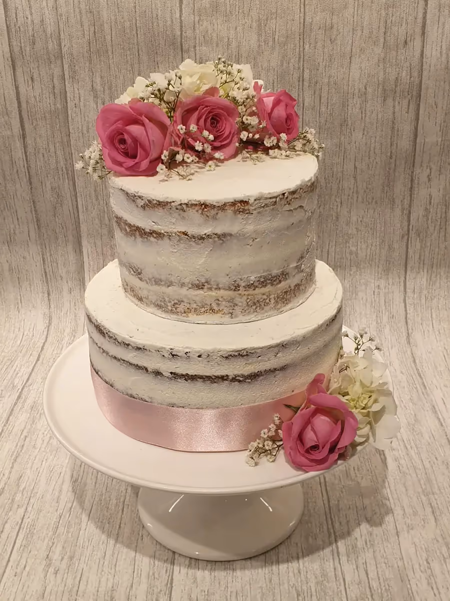 A two-tier naked cake featuring visible cake layers and thin white frosting, decorated with pink roses, white flowers, baby's breath, and a pink satin ribbon around the base.