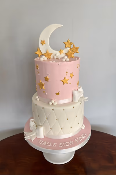 A two-tier celebration cake with a moon and stars theme. The top tier is pink, decorated with gold stars and white pearls, topped with a large white crescent moon and more gold stars. The bottom tier is white with a quilted pattern, adorned with small gold beads and white fondant bows. The cake sits on a pink board with the name 'HALLE SIEW FENG' written in white.