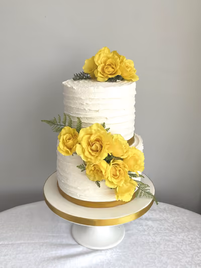 A two-tier white cake with textured buttercream frosting, adorned with bright yellow roses, green fern foliage, and gold ribbon accents, presented on a white cake stand.