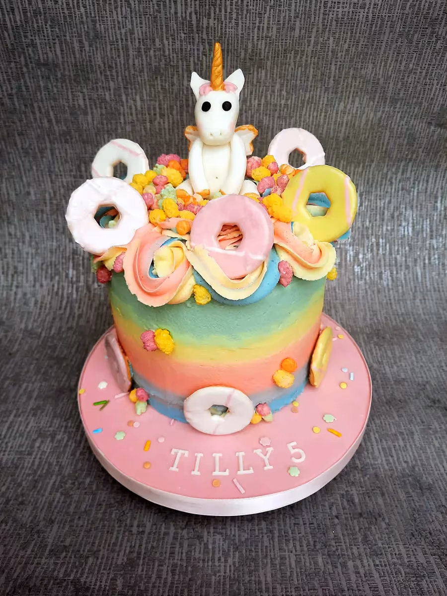 A vibrant rainbow-tiered unicorn cake decorated with mini pastel-colored donuts, colorful sprinkles, and a white fondant unicorn on top, with 'TILLY 5' written on the pink cake board.
