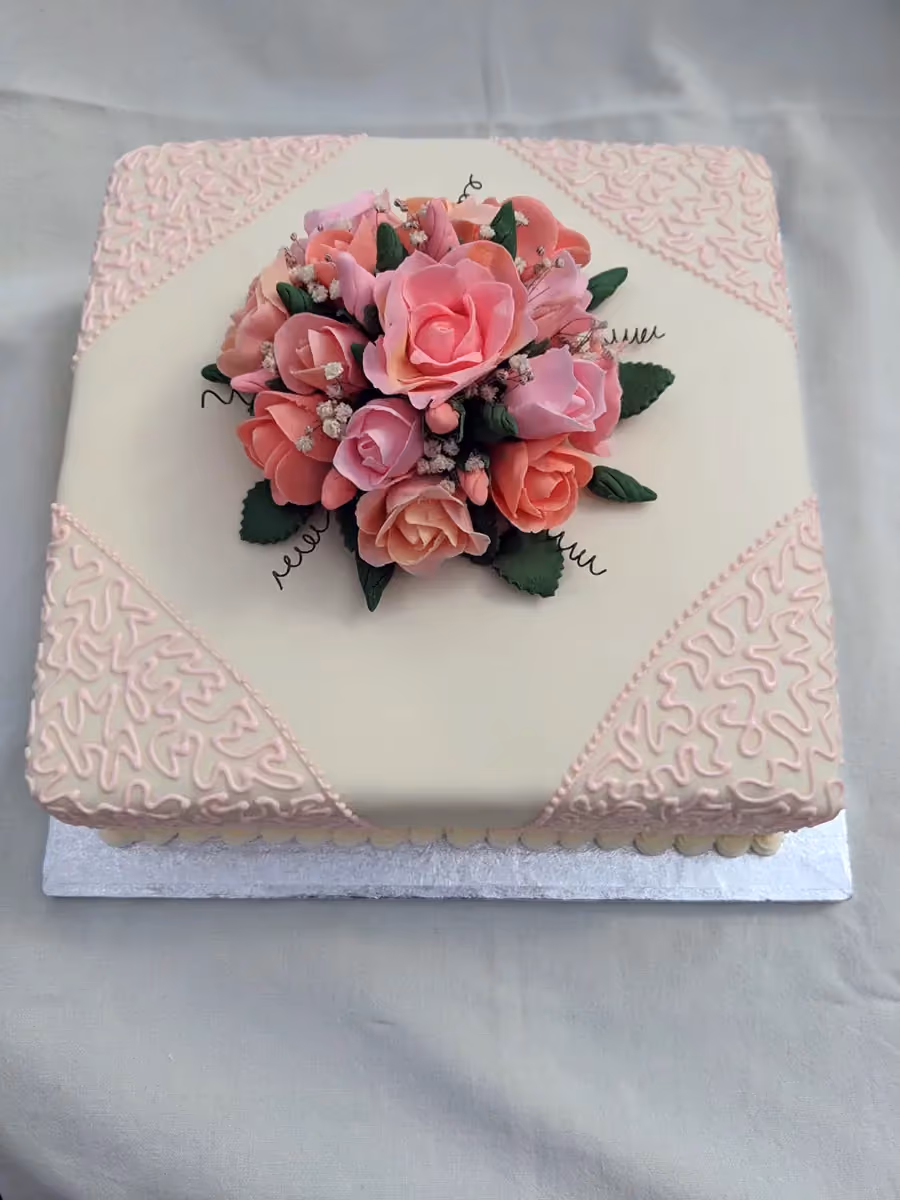 A square white fondant cake adorned with a central bouquet of pink and coral sugar roses, accented by delicate pink piped lace patterns on the corners.