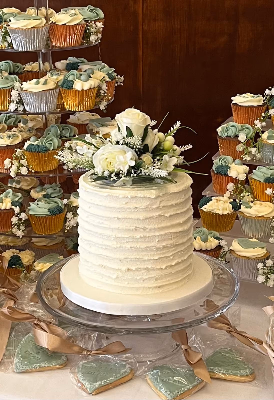 A white textured single-tier cake topped with white roses and green foliage, surrounded by tiered displays of cupcakes with green and cream frosting in metallic wrappers, and individually wrapped heart-shaped green iced cookies with ribbons.