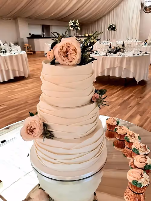 A four-tier wedding cake with white draped fondant, decorated with light pink roses and green thistles, displayed on a mirrored stand next to matching cupcakes in a wedding reception venue.