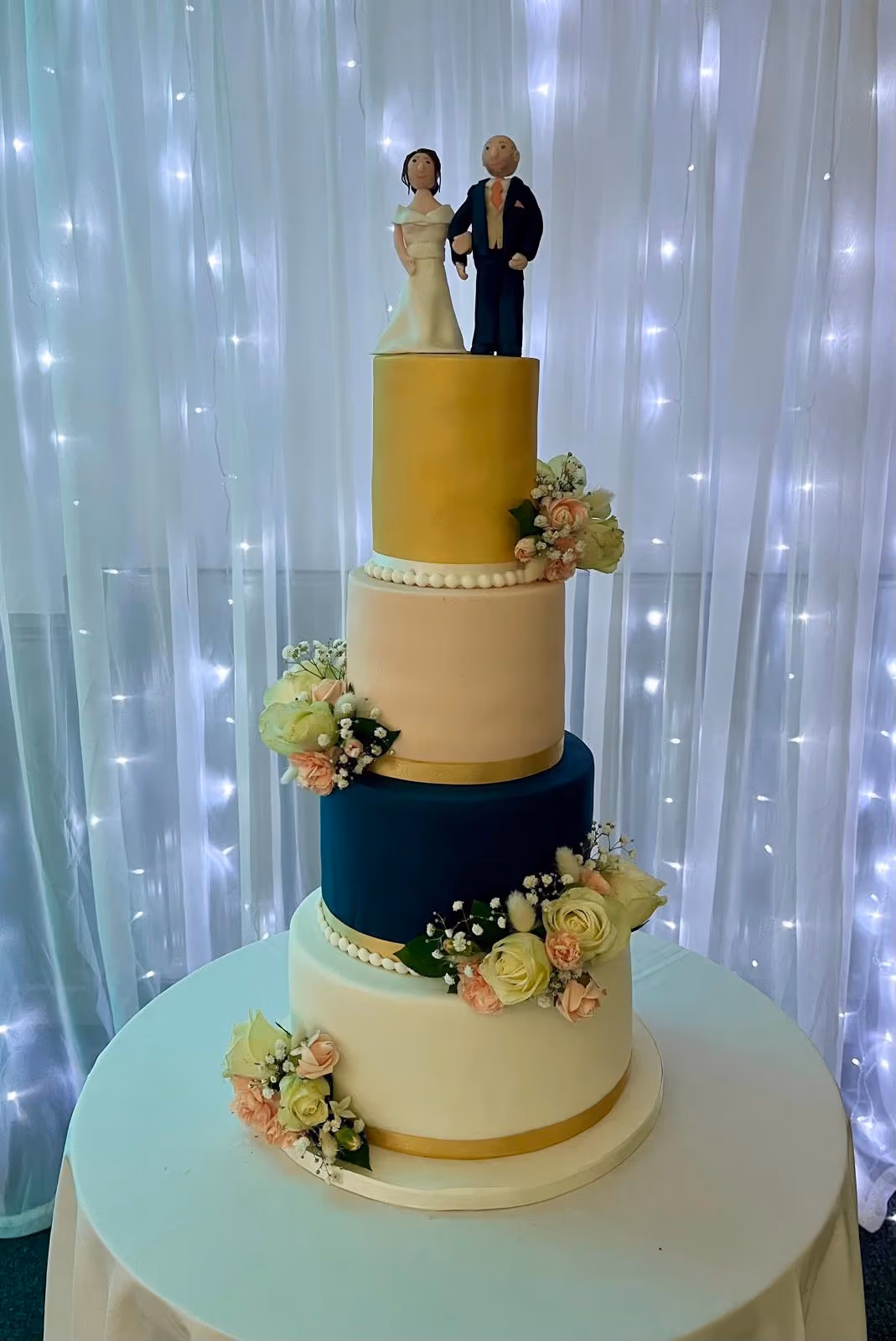 A four-tier wedding cake featuring a gold top tier, followed by a light peach, dark teal, and white bottom tier, all decorated with pearl borders, gold ribbons, and clusters of light pink and cream flowers. Bride and groom figurines top the cake.
