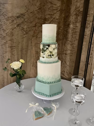 A four-tier wedding cake in white and sage green, with watercolour-effect tiers, a sage green hexagonal base tier and piped sage borders. A clear acrylic floating tier is filled with white roses and calla lilies. It stands on a white-clothed table beside a vase of white roses, floating candles and two heart-shaped favour biscuits in sage green icing.