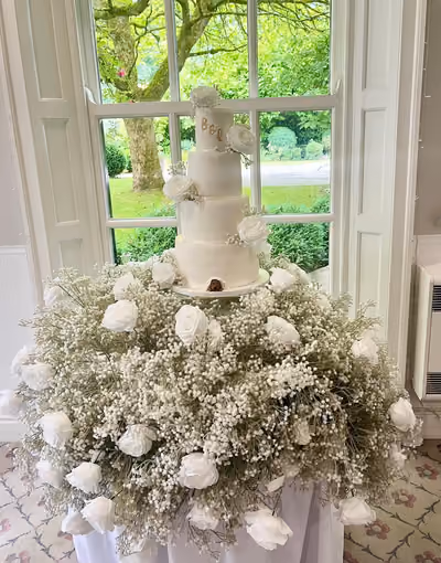 A four-tier white fondant wedding cake with gold 'B & L' initials on the top tier, white roses and baby's breath, and a small sugar dog peeking out from under the bottom tier. The cake sits in a large display of baby's breath and white roses in front of a sash window overlooking a garden.