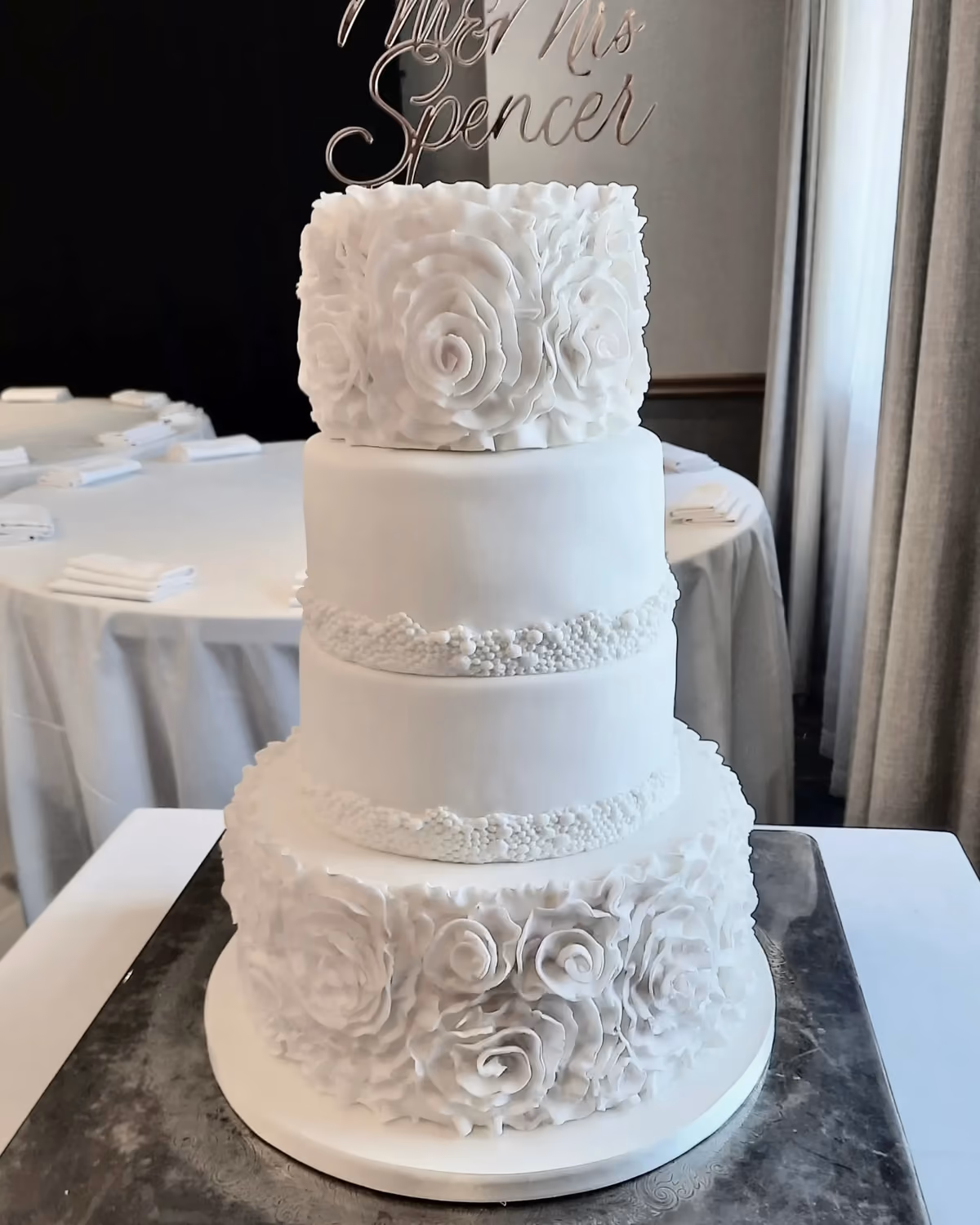 A four-tier white wedding cake with sculpted white roses on the top and bottom tiers, and bands of white pearls on the two middle tiers, topped with a silver 'Mr & Mrs Spencer' cake topper, set in a reception room.