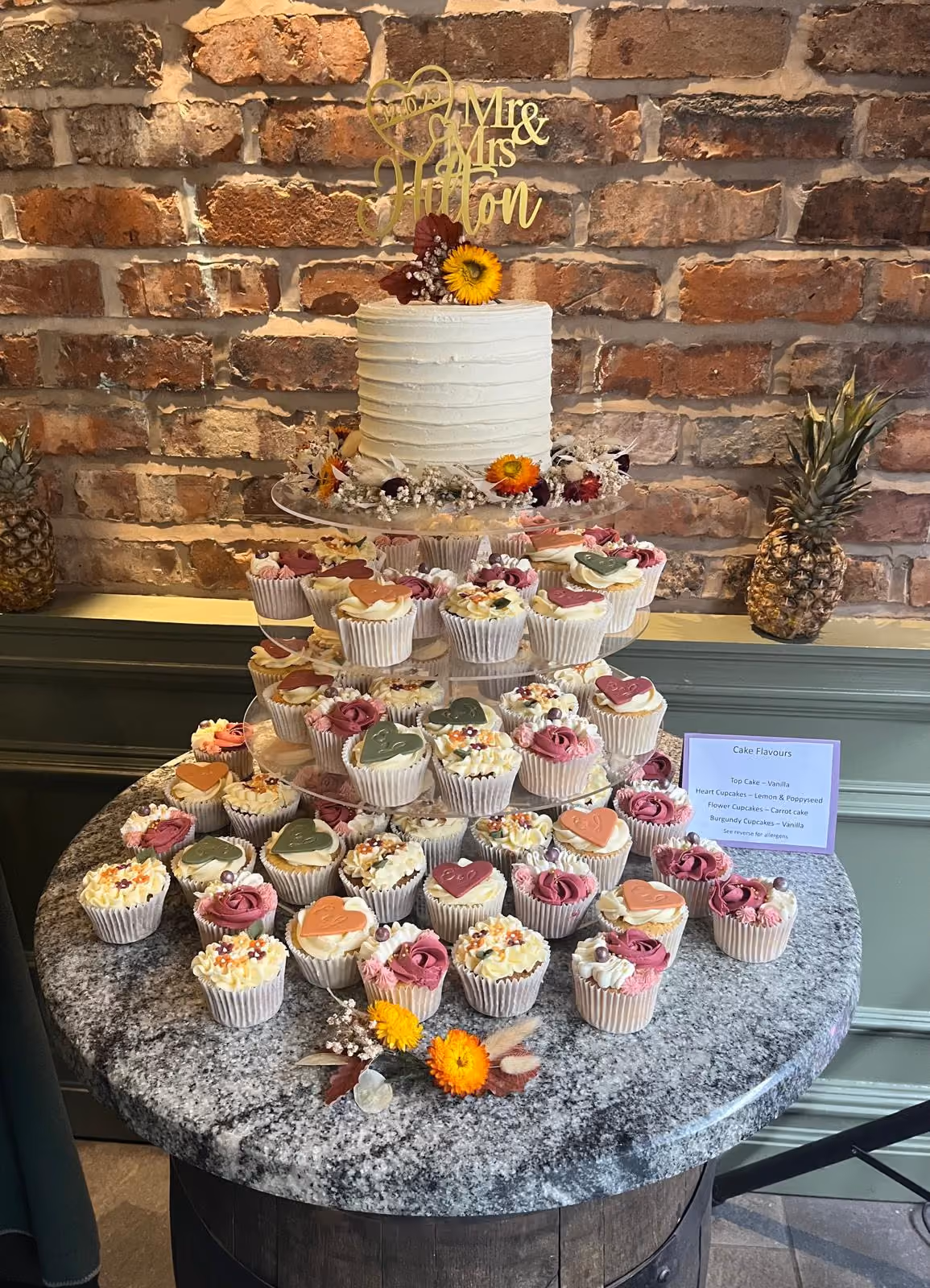A single-tier white wedding cake with a gold 'Mr & Mrs Hilton' topper and autumn floral decorations, including a sunflower, sits on a multi-tier stand above numerous cupcakes. The cupcakes feature heart and flower designs in pink, burgundy, green, and orange, with a sign indicating flavours like vanilla, lemon & poppyseed, and carrot cake.