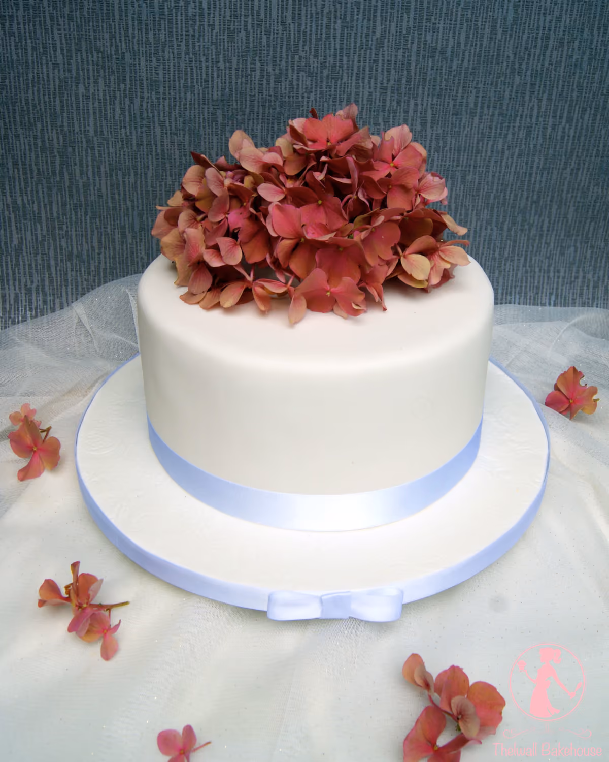 A single-tier white wedding cake, decorated with a large cluster of pink hydrangeas on top and a light blue ribbon around its base, sitting on an embossed white cake board.