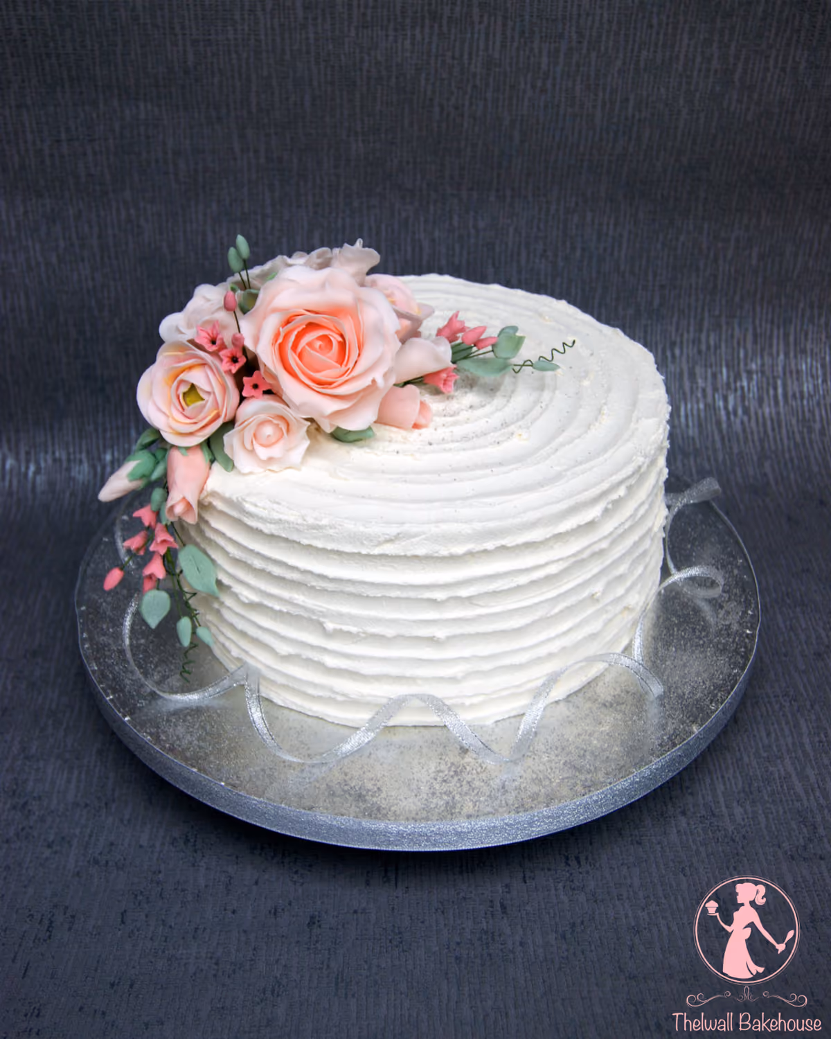 A round white cake with textured buttercream frosting, adorned with a cluster of pastel pink sugar roses, small pink blossoms, and green foliage, presented on a reflective silver cake board with a silver ribbon.