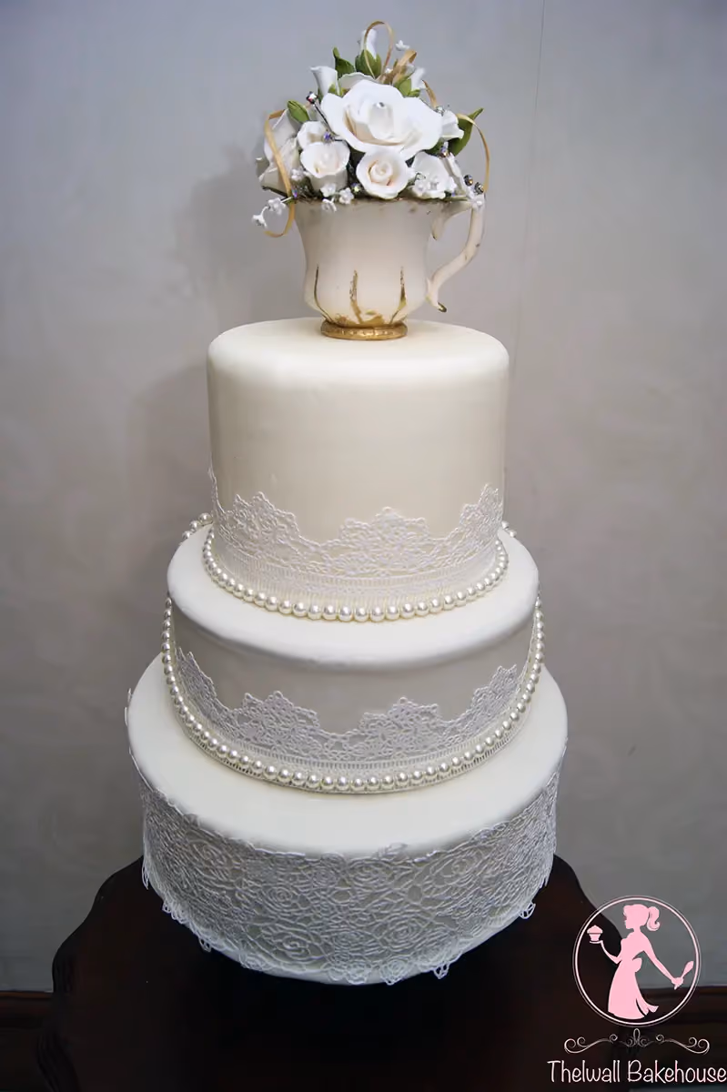 A three-tier cream wedding cake decorated with white lace patterns on each tier, pearl borders, and topped with a teacup full of white sugar roses and greenery.
