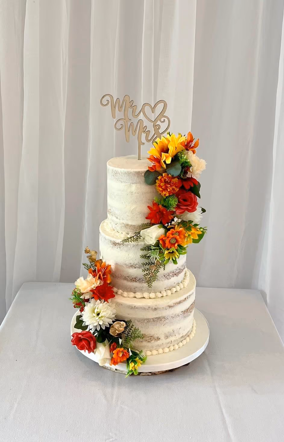 A three-tier naked wedding cake featuring a 'Mr & Mrs' cake topper and vibrant autumn-themed floral decorations, including sunflowers, red, orange, and white flowers, cascading down the sides of the tiers.
