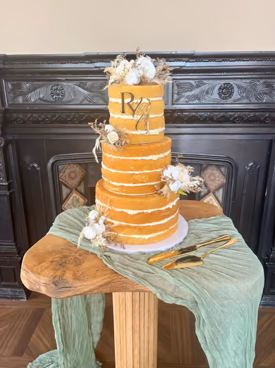 A three-tier naked wedding cake with visible sponge layers and white buttercream filling, decorated with white roses, pampas grass and dried flowers, and topped with a gold 'R & U' monogram topper. It stands on a wooden table draped with a sage green runner, beside a gold cake knife and server, in front of a black carved fireplace.