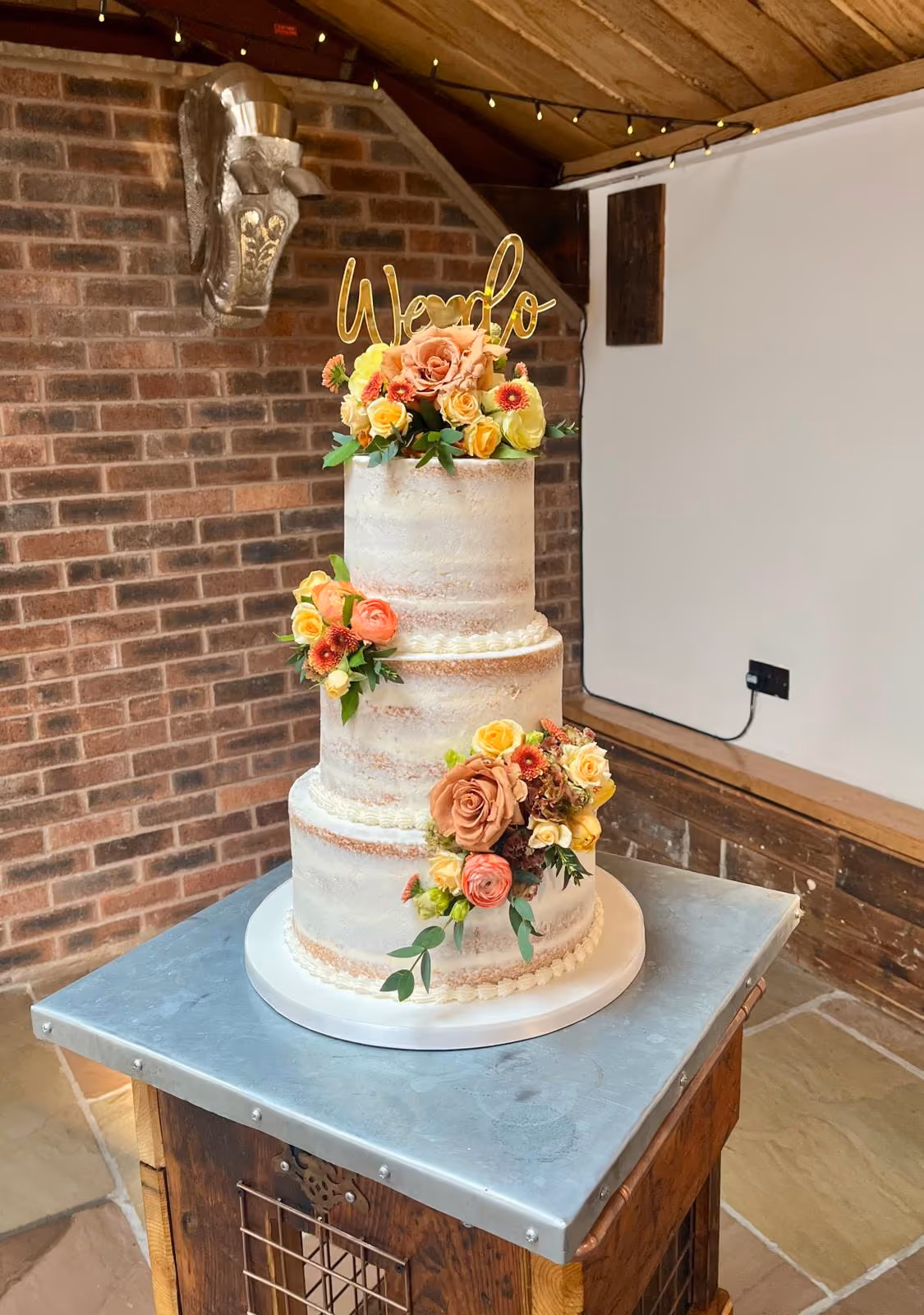 Three-tier semi-naked wedding cake decorated with clusters of orange, peach, and yellow flowers, and topped with a gold 'We do' cake topper.