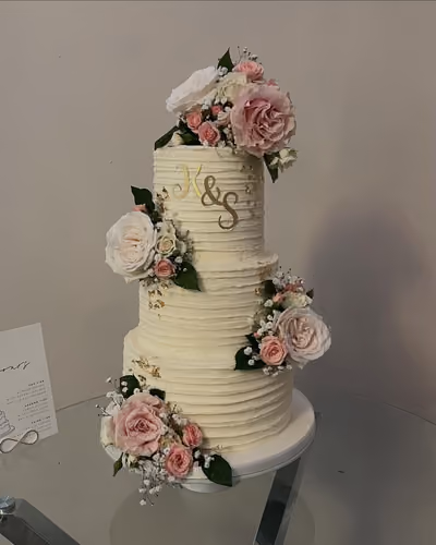 A three-tier ivory wedding cake with a horizontal textured buttercream finish, flecked with gold leaf and decorated with cascading pink and white roses, spray roses and baby's breath. A gold 'K & S' monogram sits on the top tier, and the cake stands on a glass table beside a flavours card.