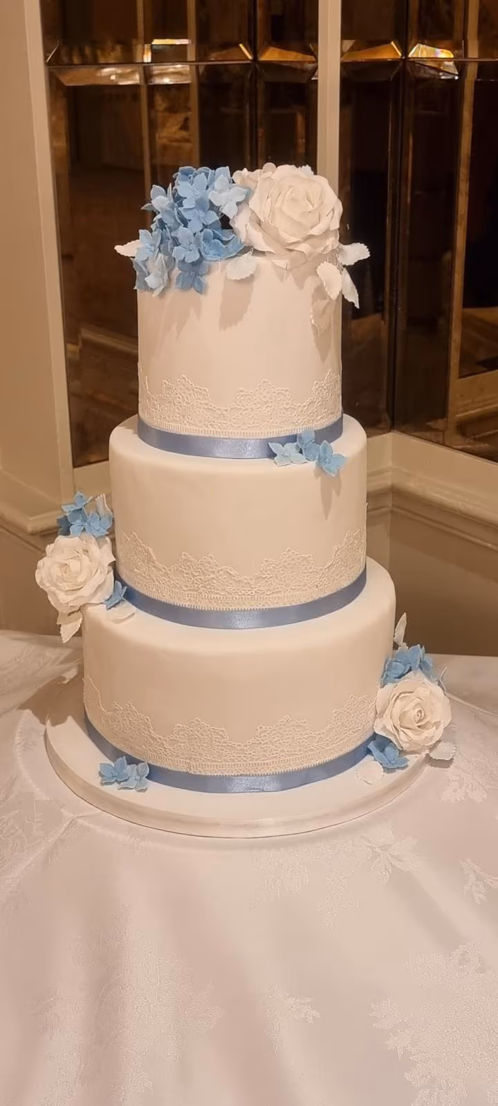 Three-tier white wedding cake with blue ribbons, lace, hydrangeas and roses