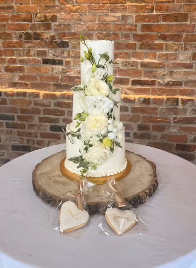 A tall three-tier white wedding cake with raw-edged tiers and a cascade of fresh white roses, lisianthus, orchids and variegated greenery. It stands on a gold board and wood slice in front of a lit brick wall, with wrapped heart-shaped 'Bride' and 'Groom' biscuits in front.