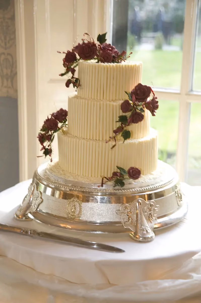 A three-tier wedding cake decorated with vertical white chocolate sticks and cascading deep red sugar roses, presented on an ornate silver cake stand with a cake knife nearby.