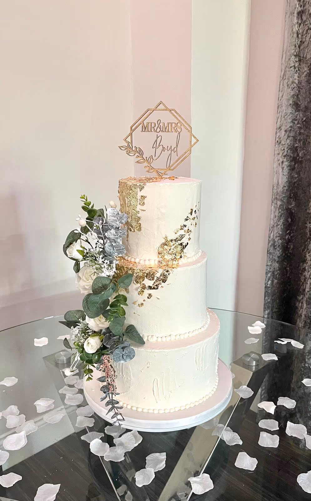 A three-tier wedding cake with white frosting and gold leaf accents, decorated with a cascading floral arrangement of white roses, green eucalyptus, and blue-grey foliage, and topped with a gold 'MR & MRS Boyd' topper.