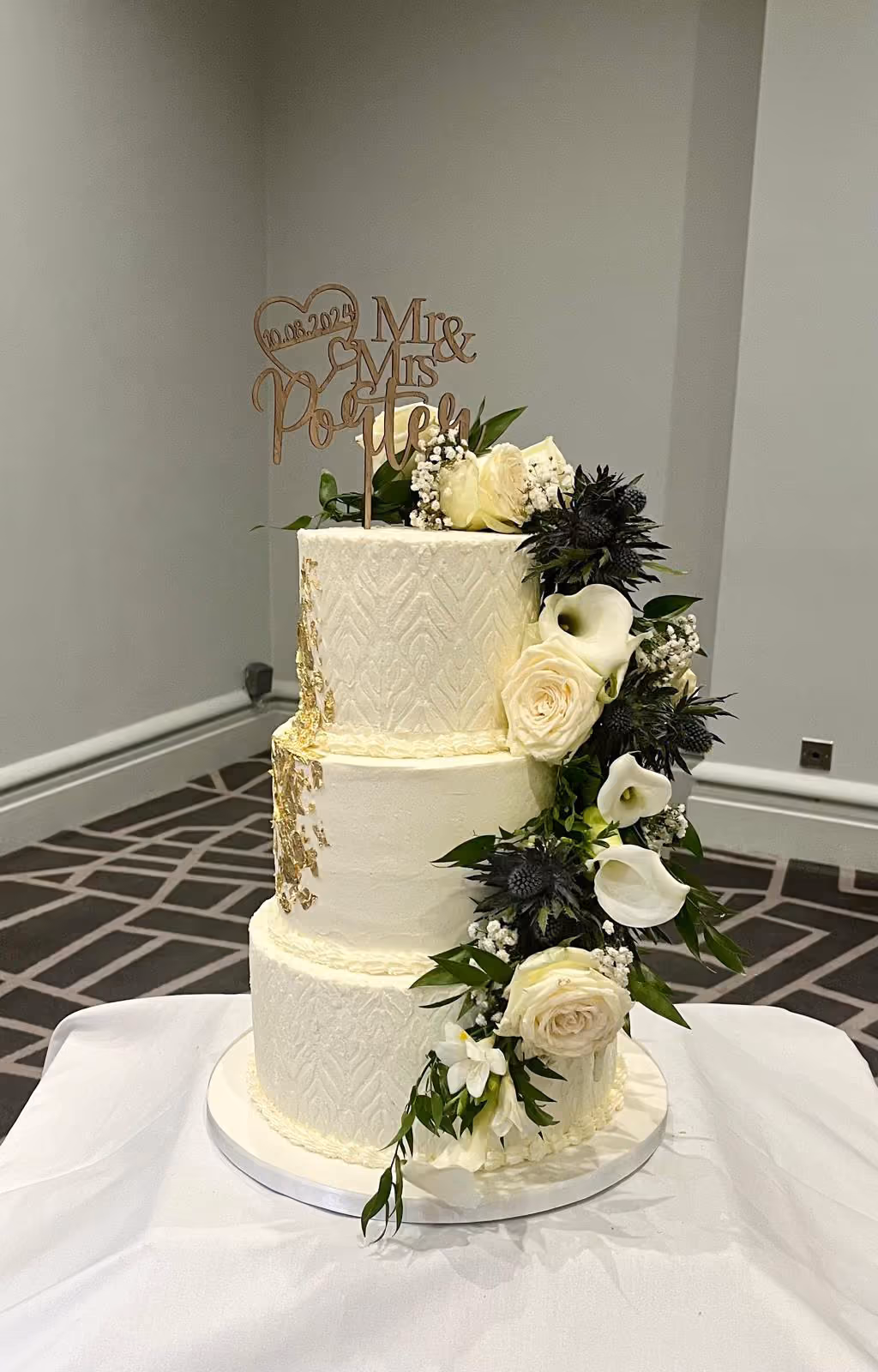Three-tier white wedding cake with gold leaf, roses, calla lilies and thistles