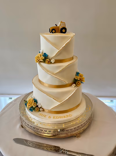 A three-tier white wedding cake with gold ribbon and trim, decorated with yellow and blue sugar flowers, and topped with a small yellow tractor. The bottom tier features the names 'SUE & EDWARD' in gold.