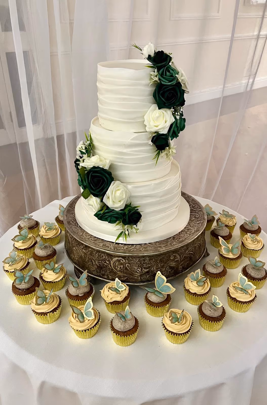 A three-tier white wedding cake with horizontal ruffled frosting, decorated with a cascade of dark green and white roses and greenery, surrounded by cupcakes with light blue butterfly toppers.