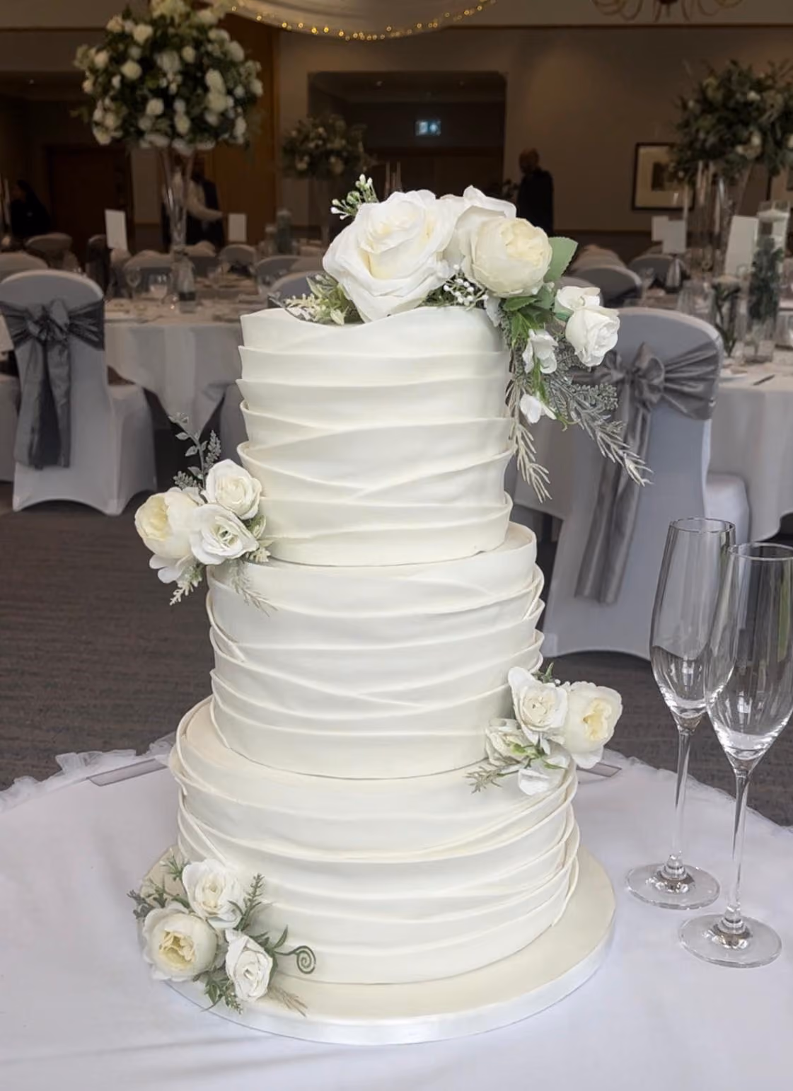 A three-tier white wedding cake with layered, ruffled fondant wraps on every tier, decorated with clusters of ivory roses, white peonies and soft greenery. It sits on a white-clothed table beside two champagne flutes, in a reception room with white chair covers, grey sashes and tall floral centrepieces.