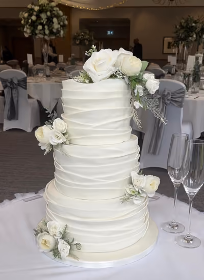 A three-tier white wedding cake with layered, ruffled fondant wraps on every tier, decorated with clusters of ivory roses, white peonies and soft greenery. It sits on a white-clothed table beside two champagne flutes, in a reception room with white chair covers, grey sashes and tall floral centrepieces.
